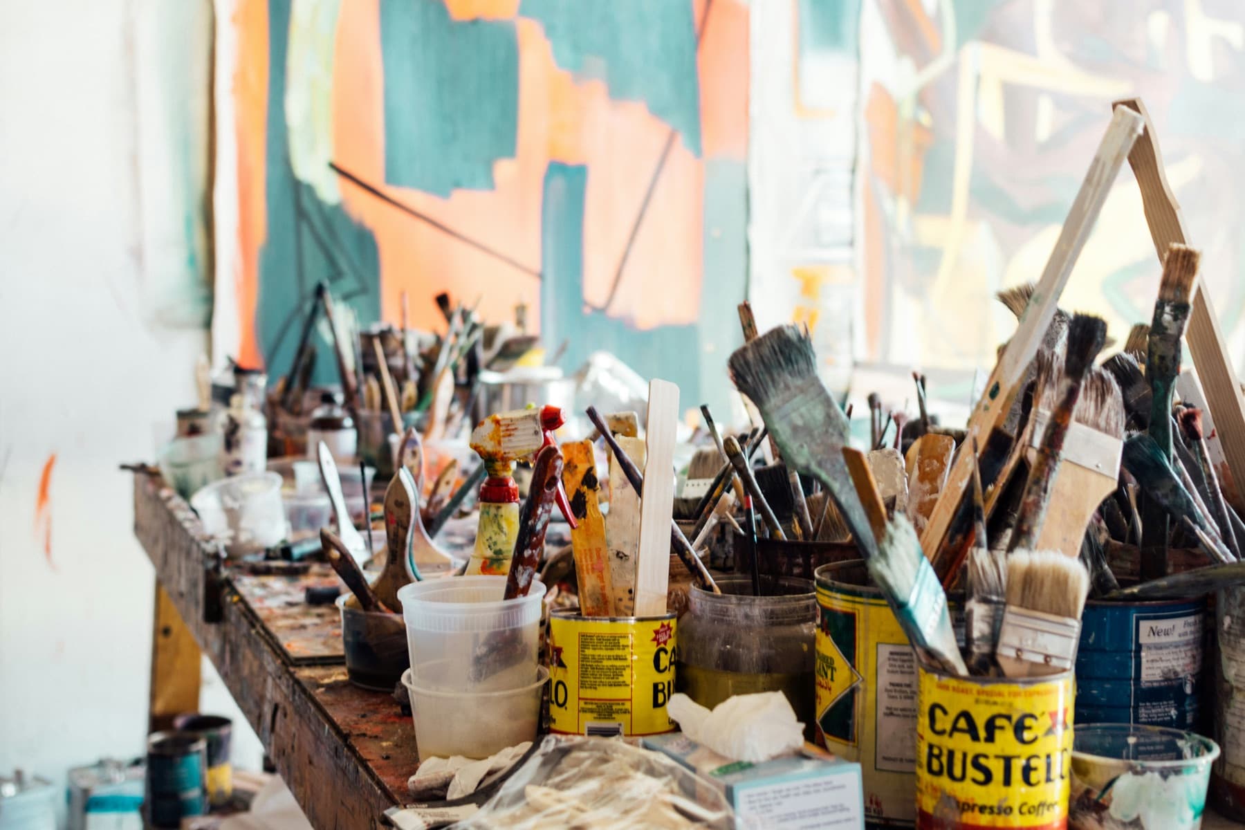 A studio bench crowded with brushes and tins in front of a half-finished painting